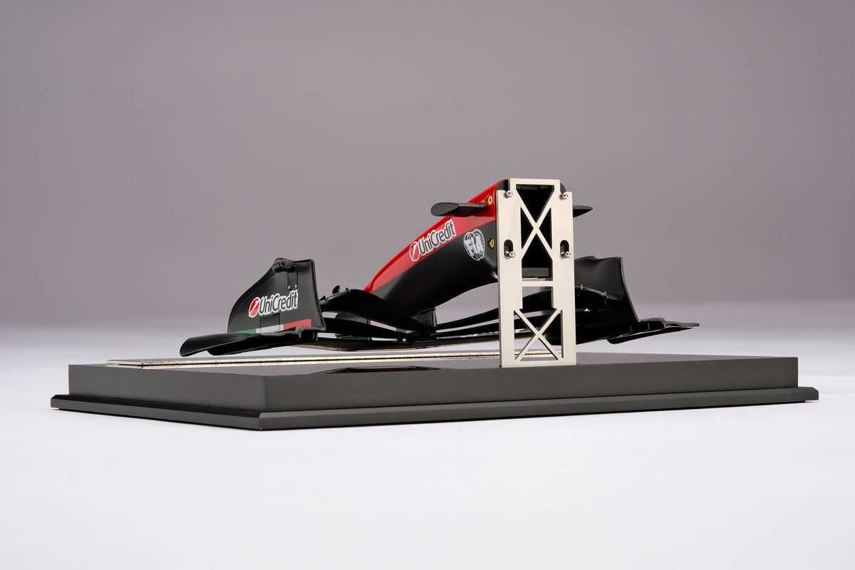 A miniature model of a red and black Formula 1 Ferrari SF-26 nosecone, mounted on a black display base against a neutral grey studio background.