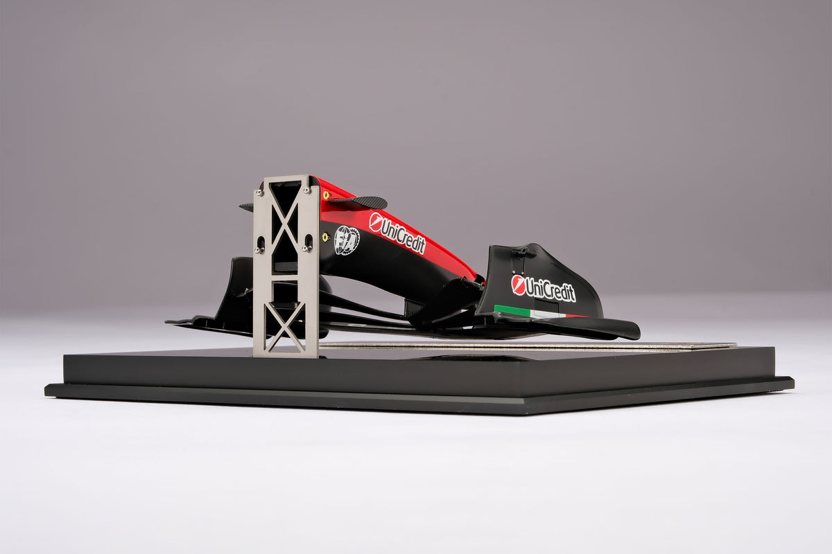 A miniature model of a red and black Formula 1 Ferrari SF-26 nosecone, mounted on a black display base against a neutral grey studio background.
