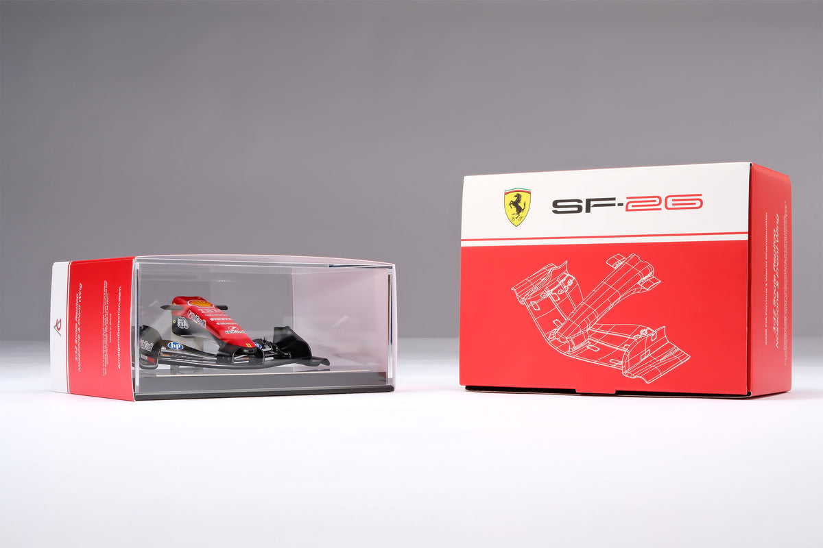 A miniature model of a red and black Formula 1 Ferrari SF-26 nosecone, mounted on a black display base surrounded by packaging, against a neutral grey studio background.
