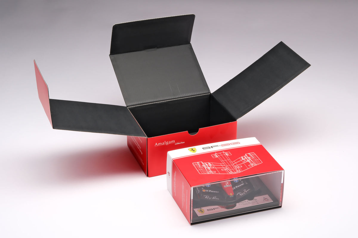 A miniature model of a red and black Formula 1 Ferrari SF-26 nosecone, mounted on a black display base surrounded by packaging, against a neutral grey studio background.