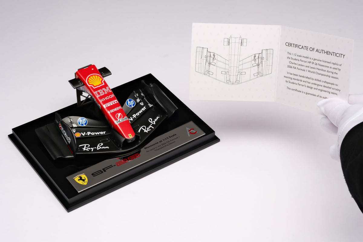 A miniature model of a red and black Formula 1 Ferrari SF-26 nosecone, mounted on a black display base surrounded by packaging, against a neutral grey studio background.