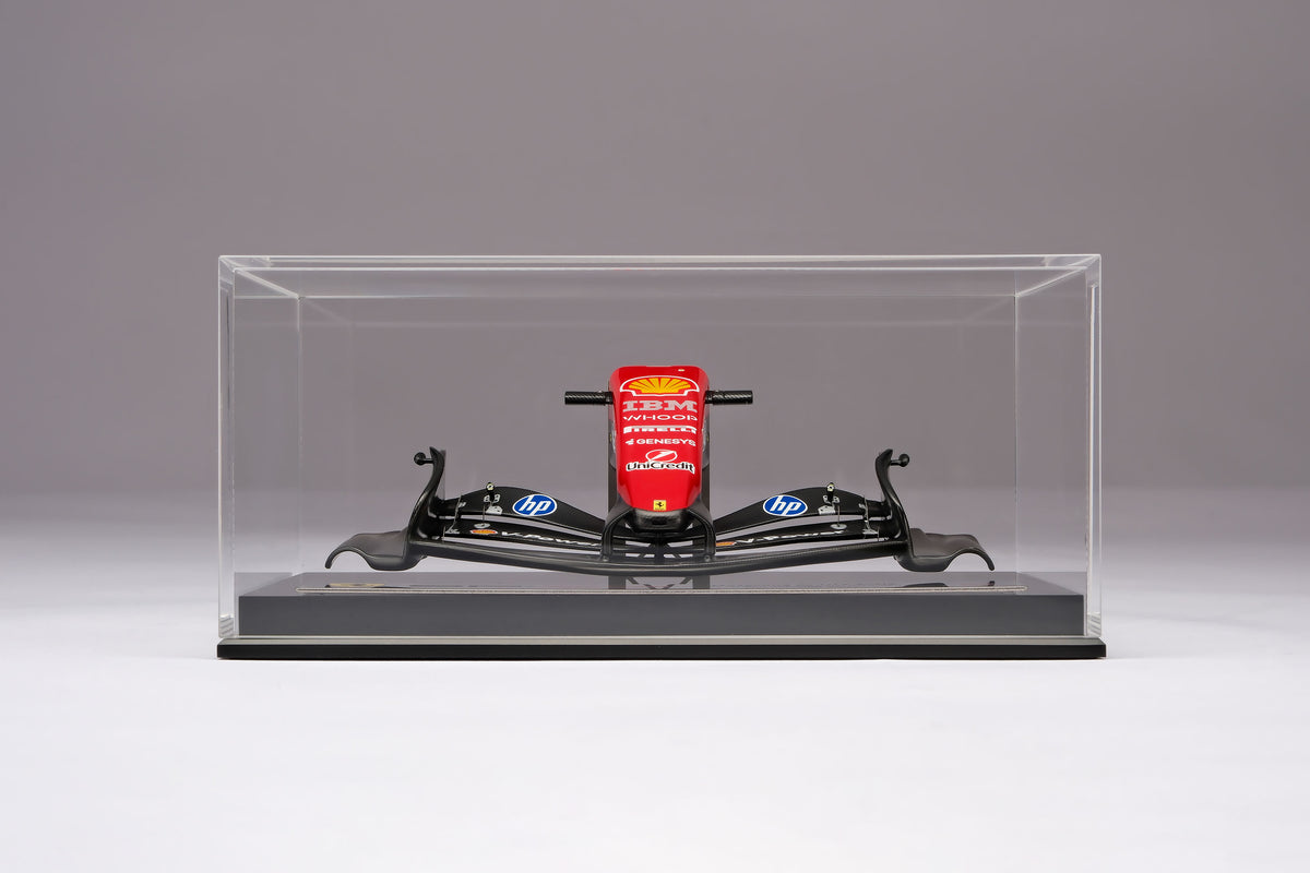 A miniature model of a red and black Formula 1 Ferrari SF-26 nosecone, mounted on a black display base inside a acrylic display box, against a neutral grey studio background.