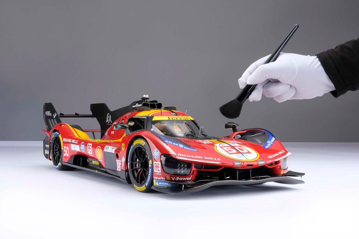 A red and yellow Ferrari 499P 2025 Le Mans racing car at 1.8 scale with multiple sponsors, displayed on a light surface and with a hand brushing the model to display the scale.