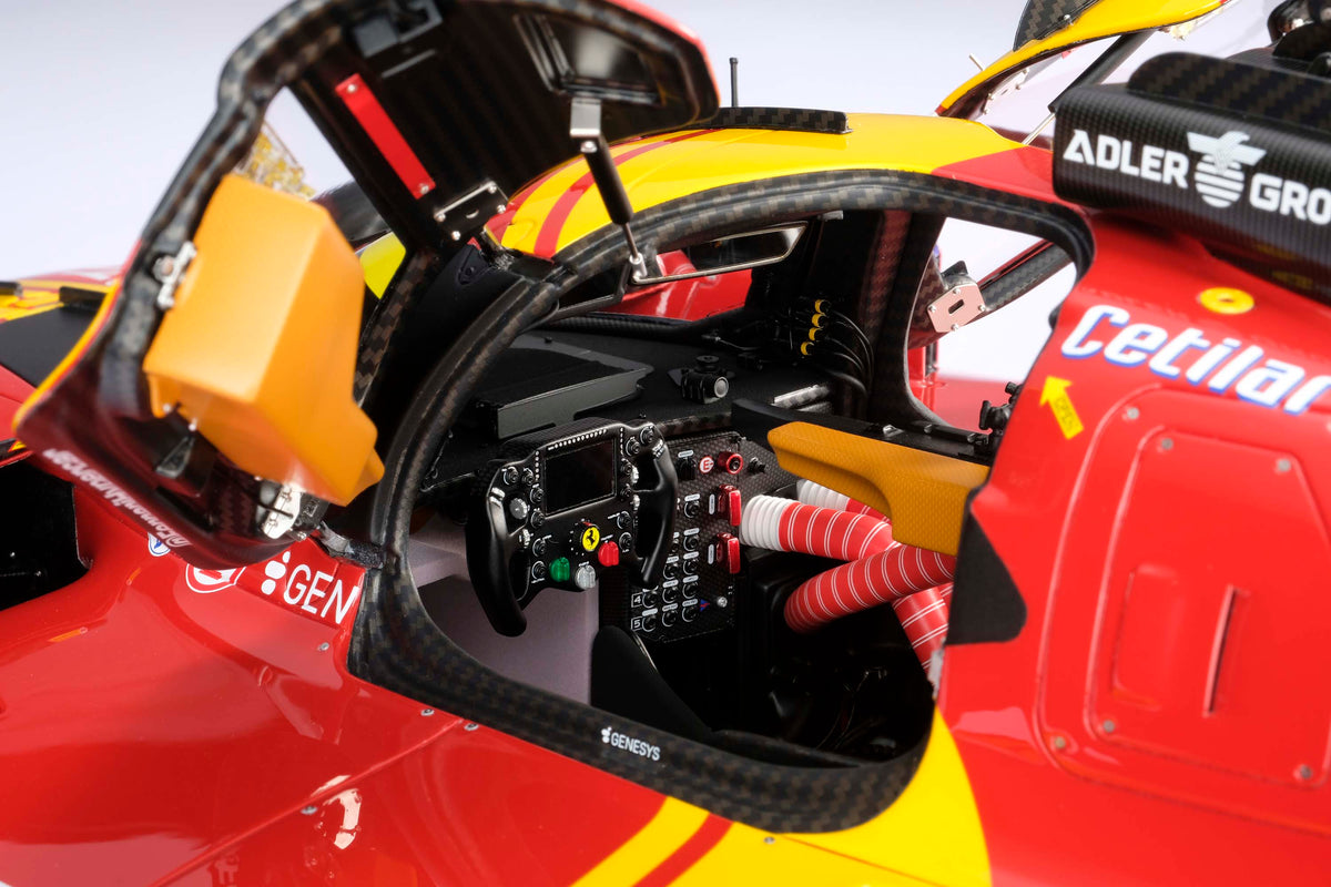 Close-up of a red and yellow Ferrari 499P cockpit interior, showing the steering wheel, instrument panel, harness, and part of the car body with sponsor logos 