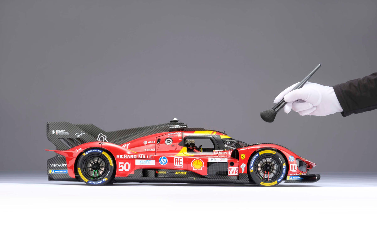 A red and yellow Ferrari 499P 2025 Le Mans racing car at 1.8 scale with multiple sponsors, displayed on a light surface and with a hand brushing the model to display the scale.