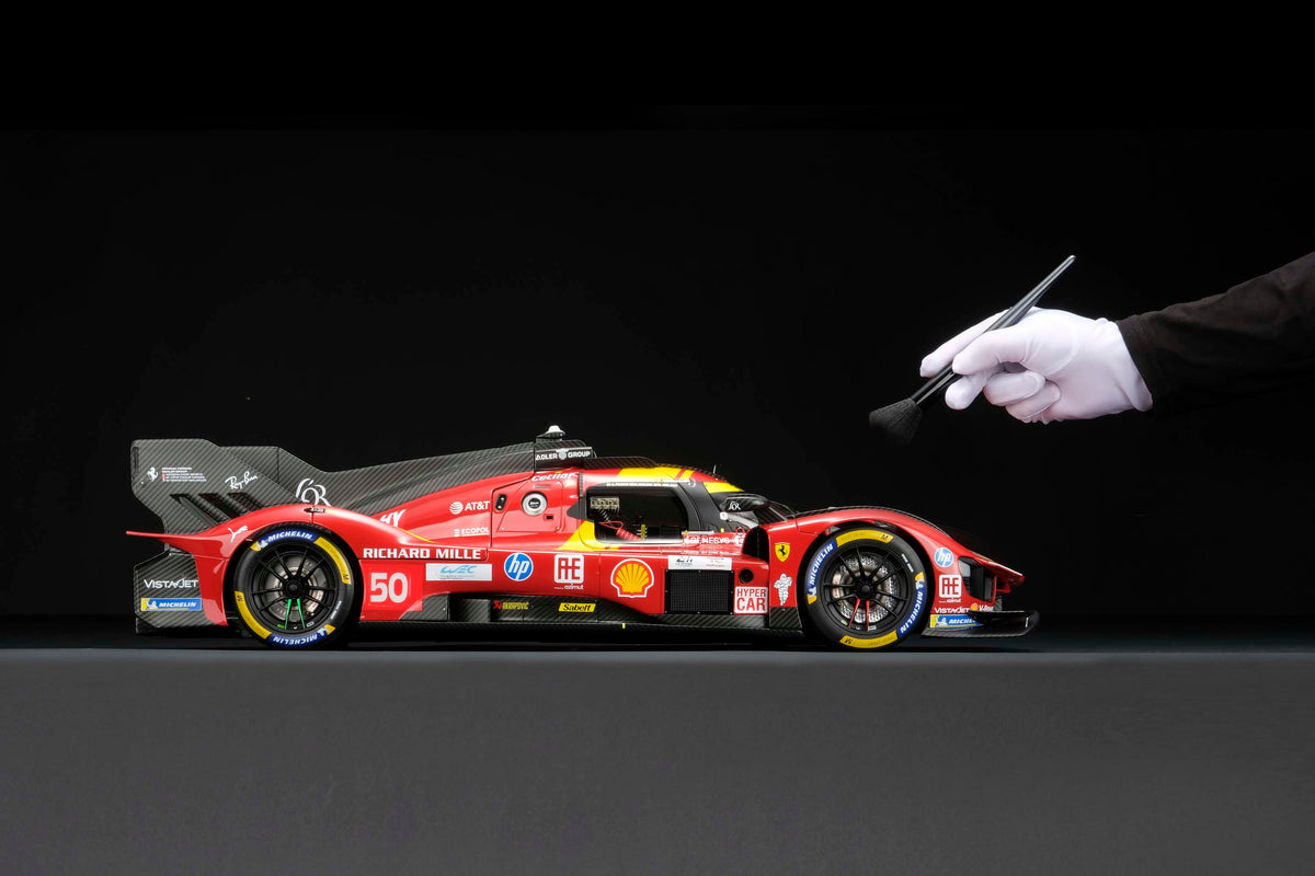 A red and yellow Ferrari 499P 2025 Le Mans racing car at 1.8 scale with multiple sponsors, displayed on a dark surface against a black background and with a hand brushing the model to display the scale.