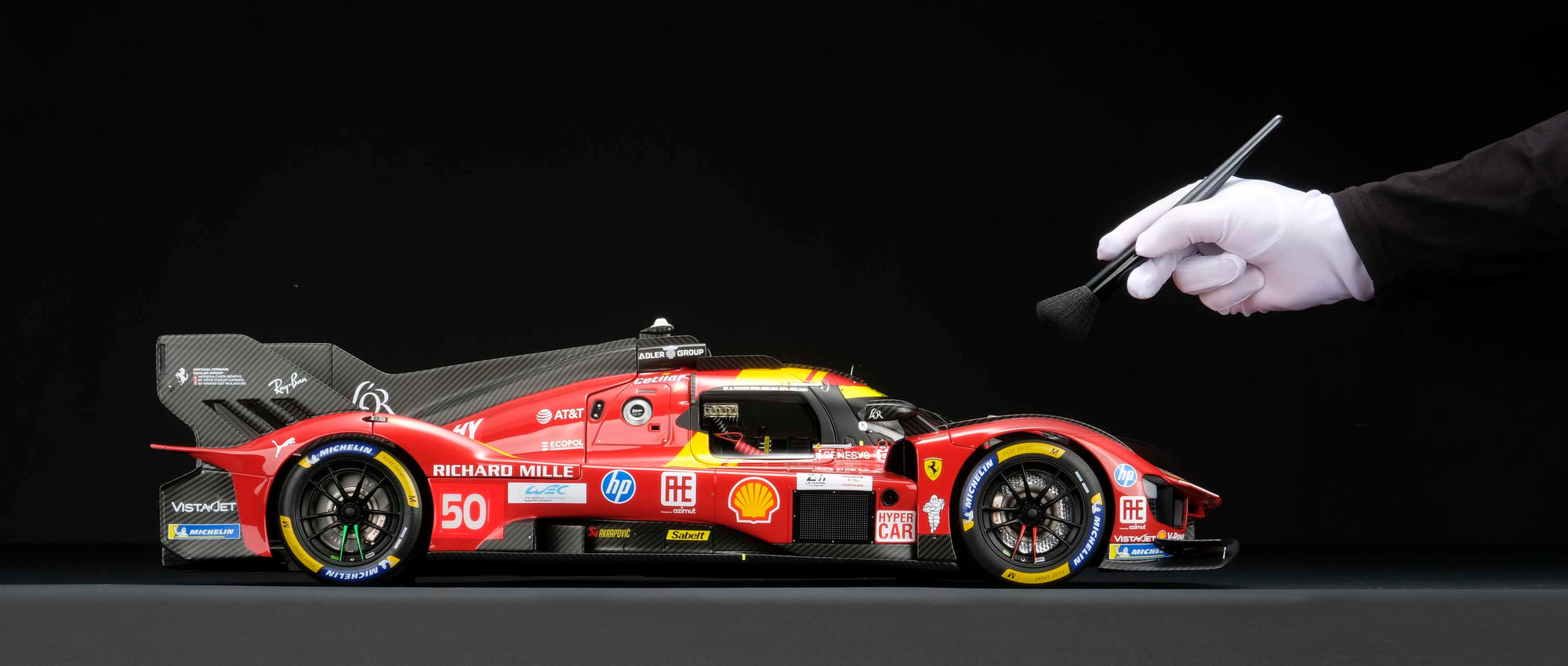 A red and yellow Ferrari 499P 2025 Le Mans racing car at 1.8 scale with multiple sponsors, displayed on a dark surface against a black background and with a hand brushing the model to display the scale.